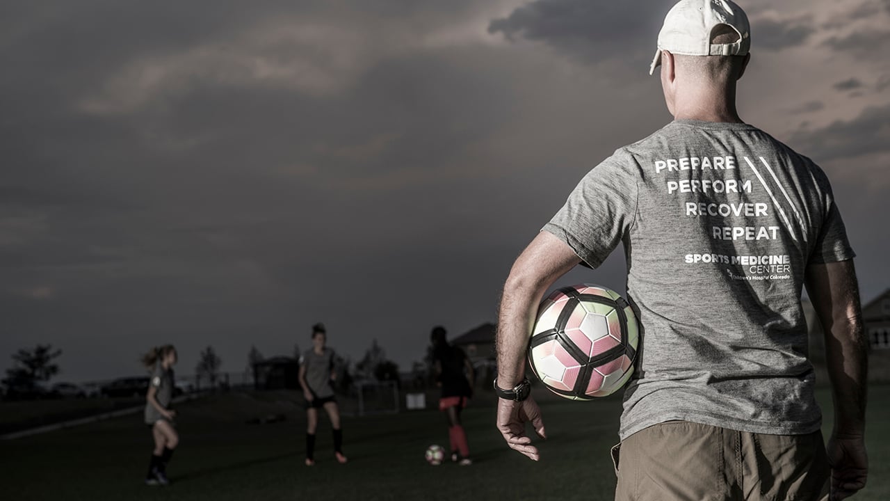 A man with a Children's Hospital sports medicine t-shirt, facing a field, holding a soccer ball