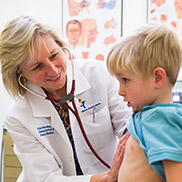 A woman with short blonde hair and a white lab coat listens to a blond boy's heart through a stethoscope.