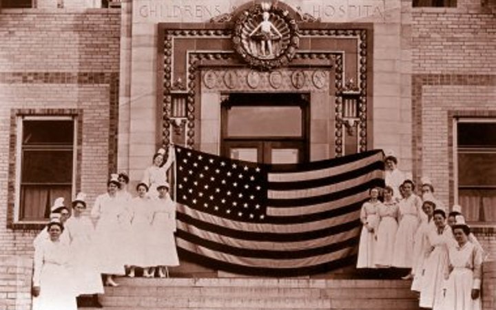 Nurses in white gowns and white hats stand on each side of the steps leading up to the hospital entrance and hold a giant American flag at the top of the stairs