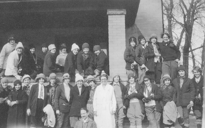 An old photo of about 30 kids gather on the porch - some sitting on the porch wall, others standing in front of it - with a woman in a white gown