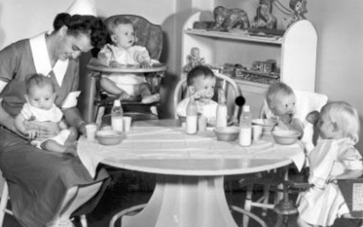 An old photo of a nurse wearing a dress and white hat holding a baby on her lap while another baby sits in a high chair and three toddlers sit around a table eating.
