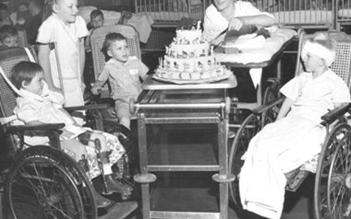 An old photo five kids, four in wheelchairs and one on crutches, sitting around a three-tier cake sitting on a cart