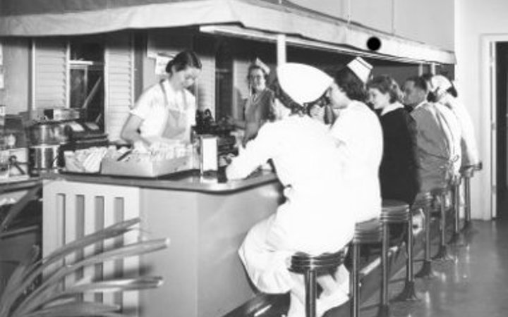 A black and white photo of two women standing under an awning and behind a dining counter serve doctors and nurses who are sitting on stools at a snack bar.