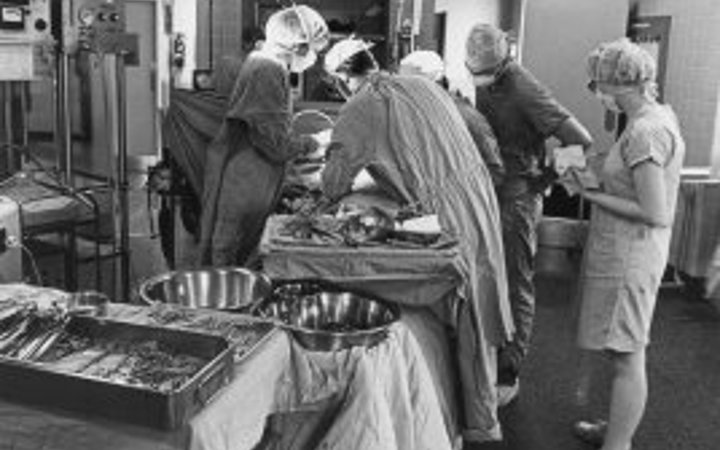 A black and white photo of an operating room with four surgeons working on a patient while a nurse looks on.