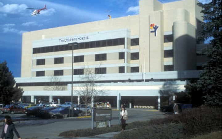 Outside view of The Children's Hospital with three rows of windows and the balloon boy logo on the building while a helicopter flies away in the bright blue sky.
