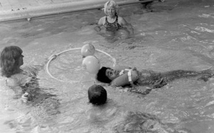 A black and white photo of two woman standing in a pool with balls and a hula hoop while two boys float in the water, one on his stomach with his head up and the other on his back wearing a life vest.