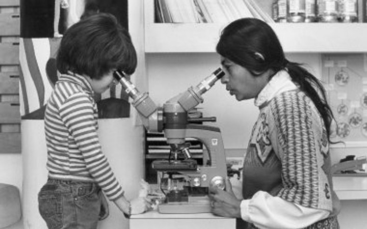 Black and white photo of a woman (Dr. Taru Hays) with long dark hair looking into a microscope with two viewfinders. A young kid wearing a striped shirt looks into the other viewfinder on the other side of the microscope.