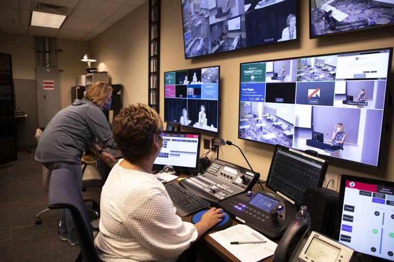 Two women working in front of four large mounted monitoring screens showing security camera footage and four computers like a command center.