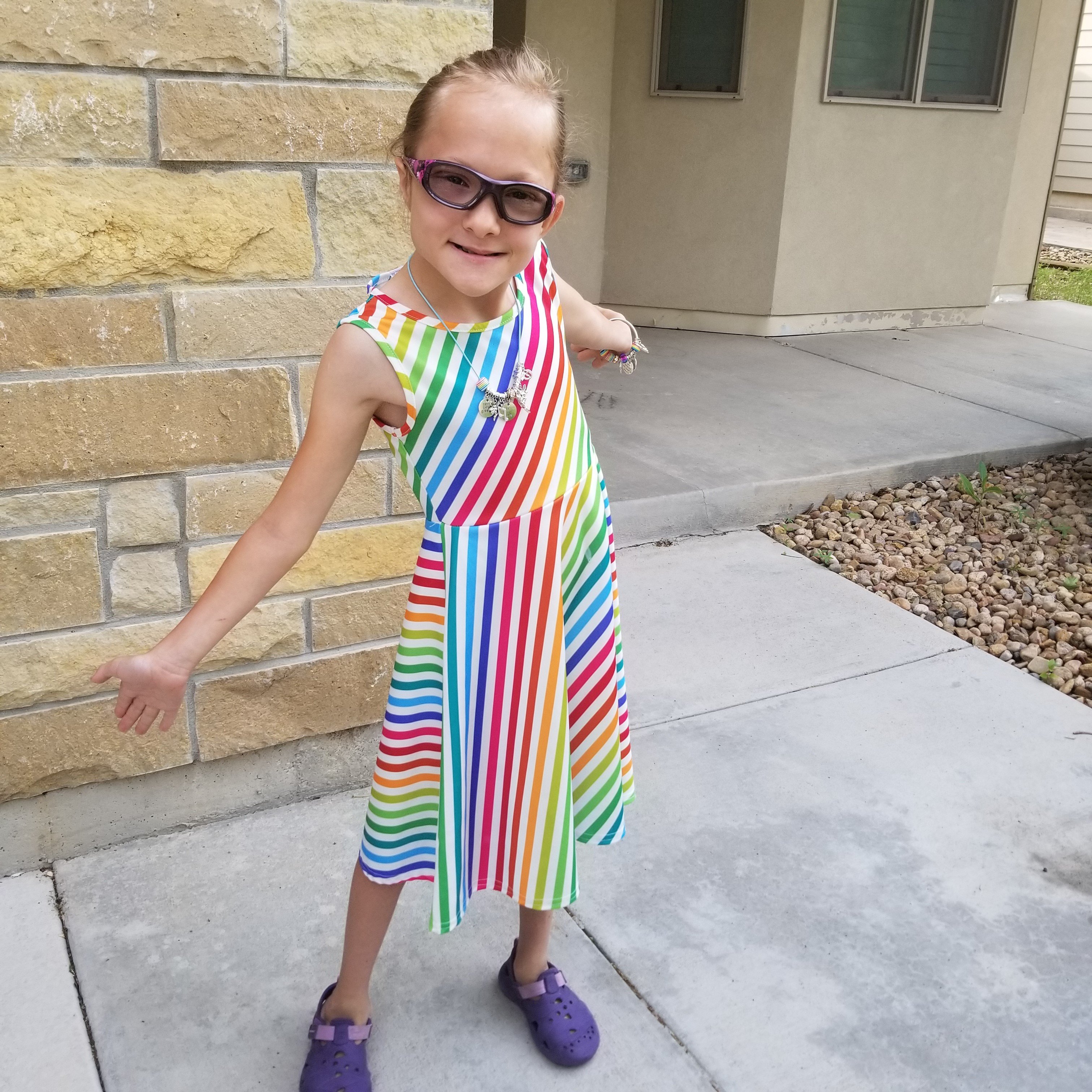 A girl (Eve) in a rainbow dress with her arms outstretched, wearing protective glasses for pontine tegmental cap dysplasia 