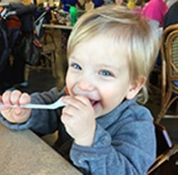 blonde boy in a restaurant with a spoon in his mouth smiling. 