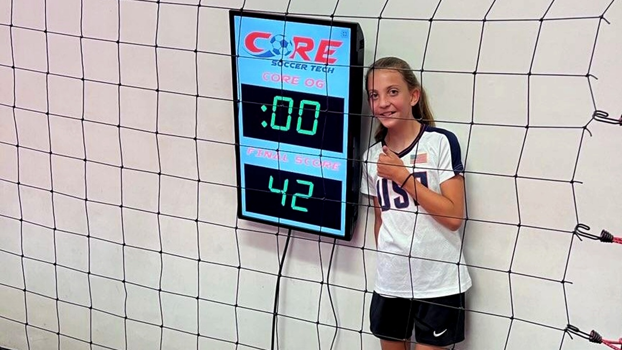 Kendall smiles with a thumbs up next to a soccer scoreboard.