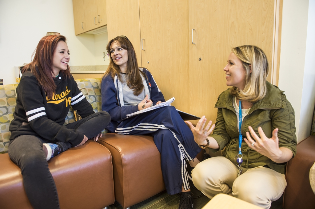 A blonde woman wearing yellow pants and a green jacket squats next to two ladies sitting in brown chairs while they have a conversation. They both have long brown hair. One is wearing a black sweatshirt with Colorado in yellow lettering and the other is wearing a blue track suit and taking notes in a notebook.