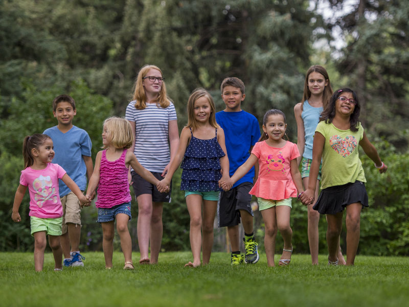 A group of 9 kids - five in front and four in back - hold hands and walk in a grassy field with trees behind them. The kids are ages 4-15 and wearing brightly colored clothes in a variety of colors.