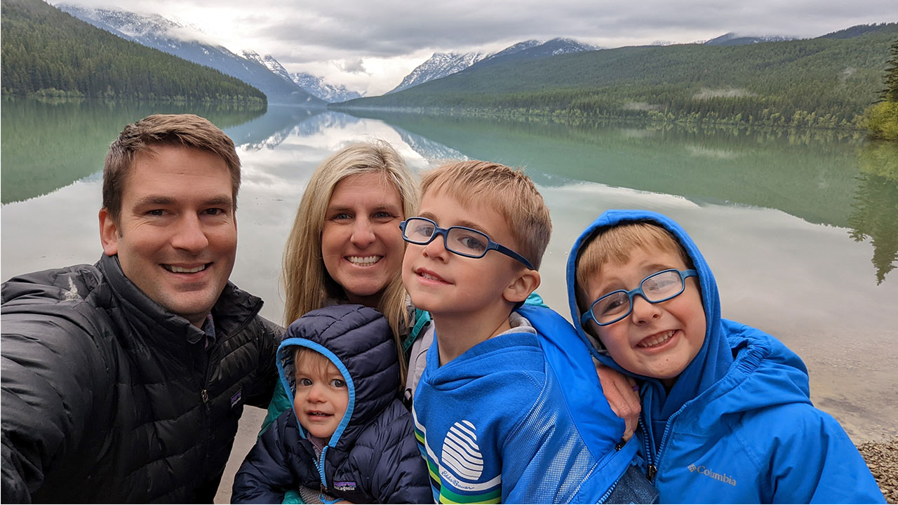 A family of a man, woman, two boys and a girl smile in front of a lake and mountains.