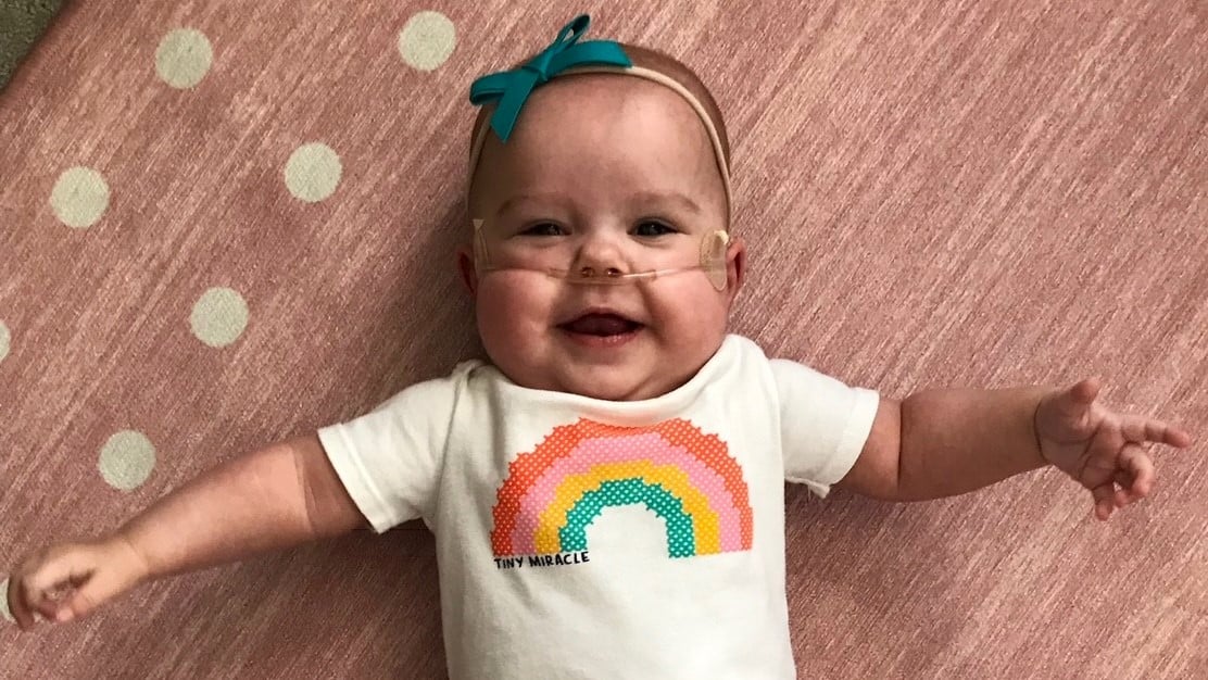 Emma, as an infant, wears a bow on her head and a shirt with a rainbow on it that says “Tiny Miracle.”
