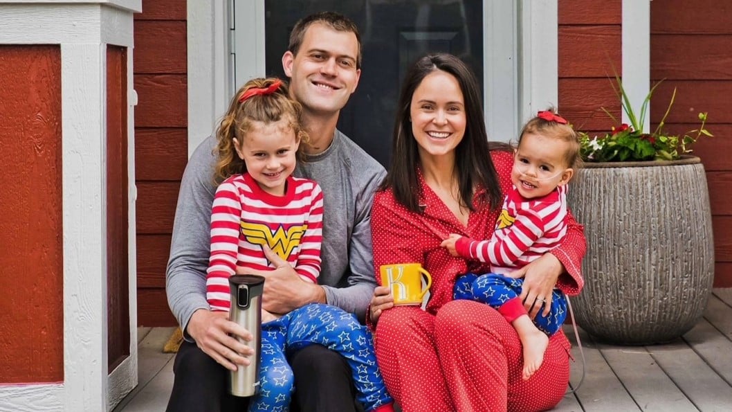 Emma's family of four sits outside in pajamas and the two young girls wear matching Wonder Woman pajamas.