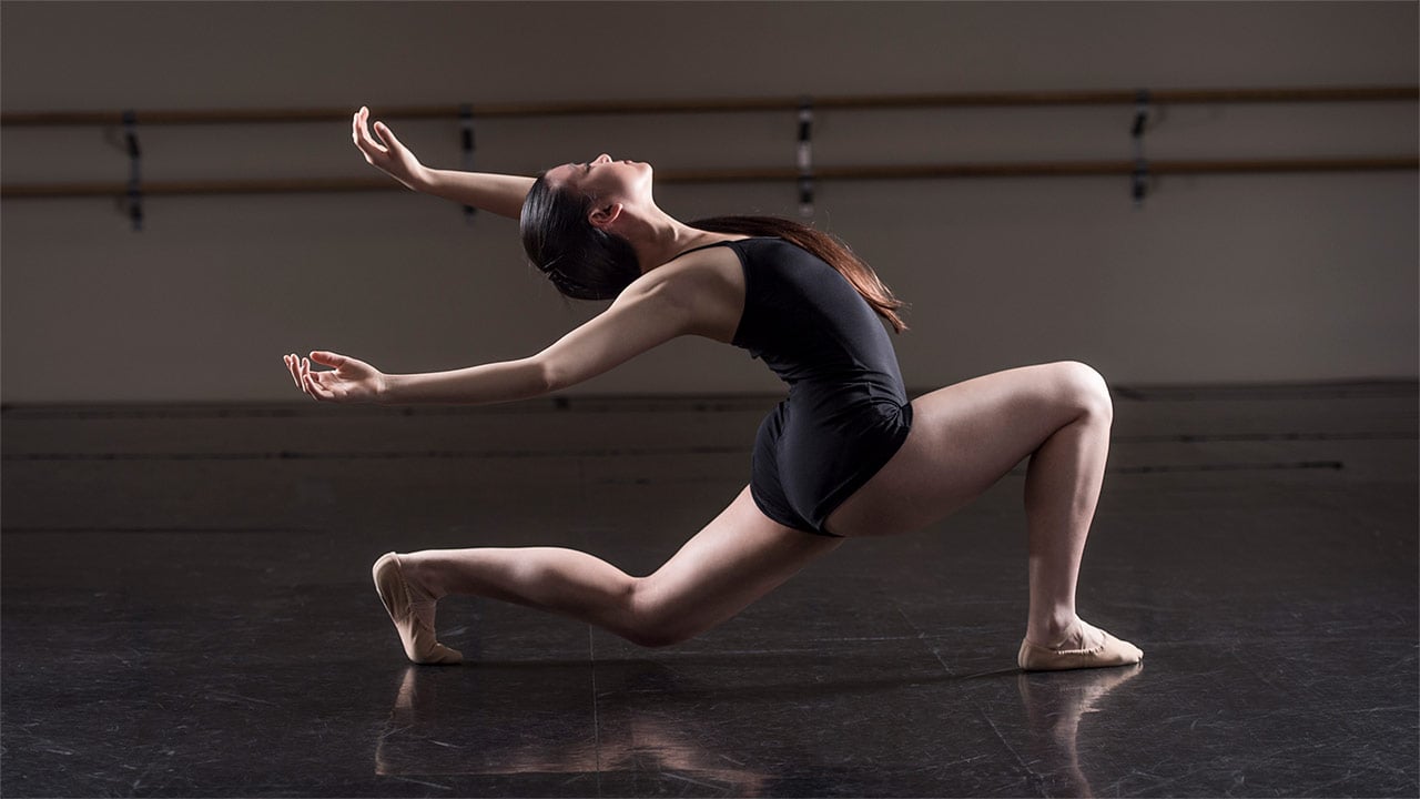 A teenage ballerina in a black leotard bends back low to the ground in a dance studio.