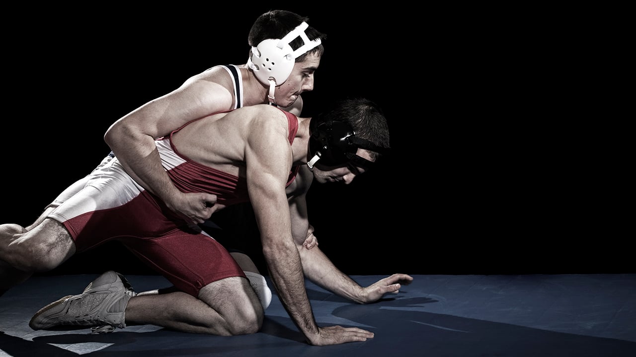 A boy wearing a red wrestling uniform and black head gear is on his hands and knees while another wrestler wearing white head gear reaches over him.