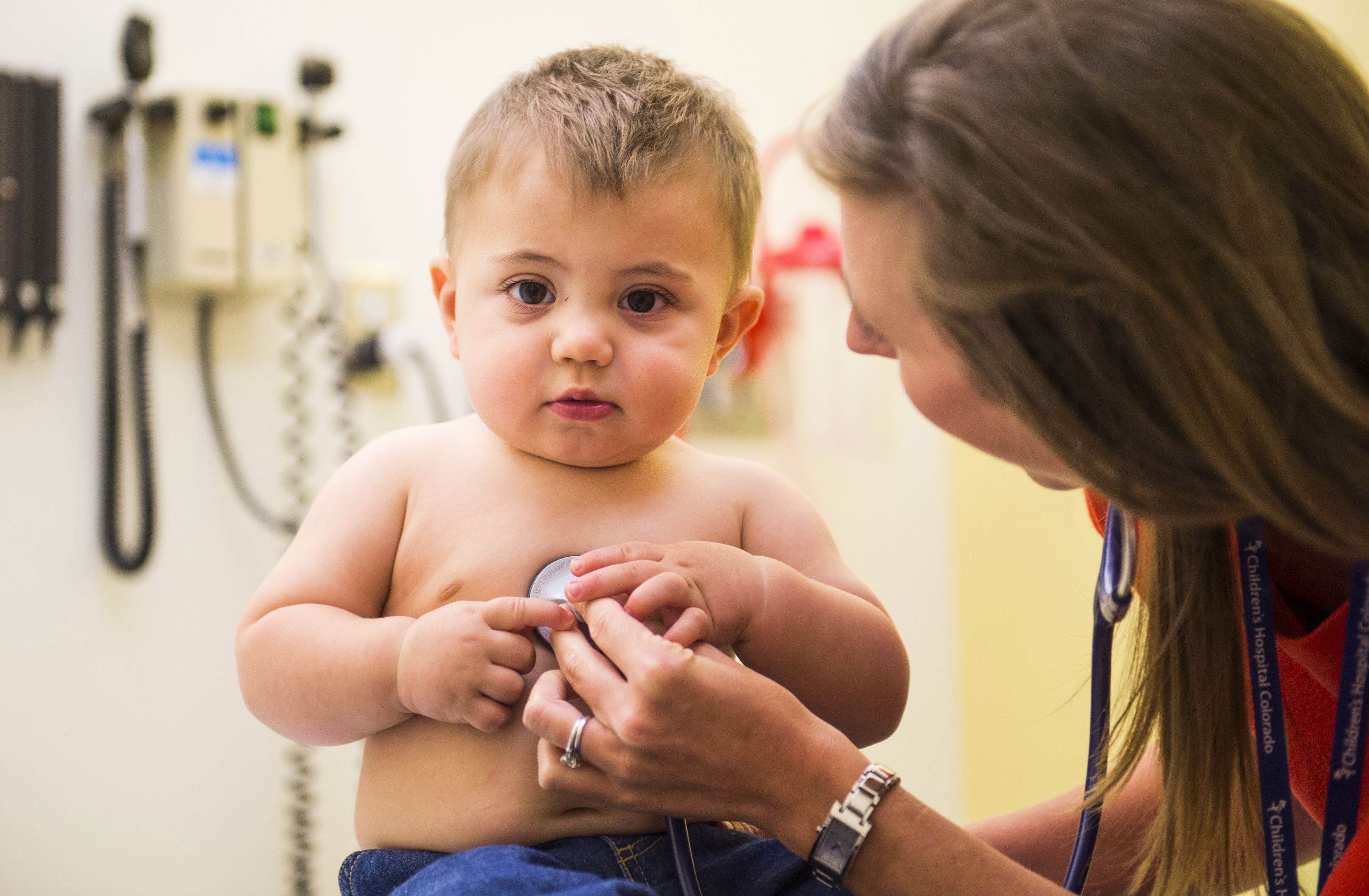 A toddler with brown hair sits in a doctor's office while a doctor examines him with a stethoscope.