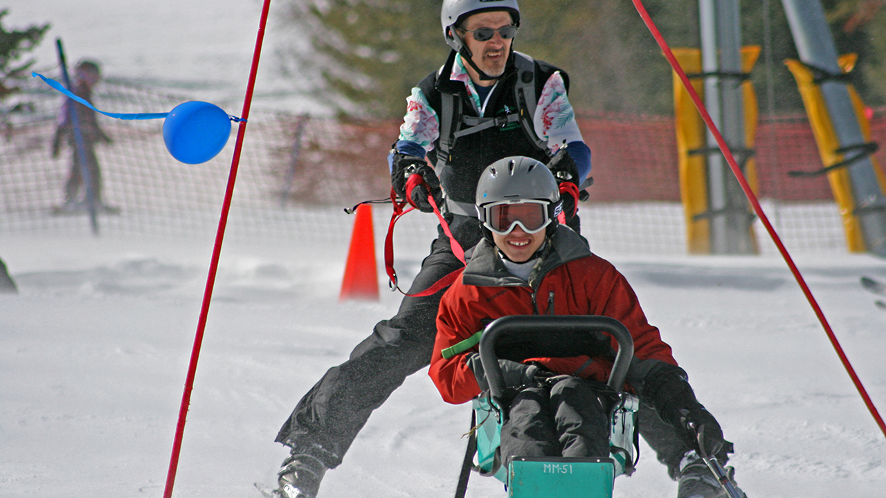 A kid in a red jacket, gray helmet and goggles rides in a green ski sled while a man in a black ski suit, gray helmet and sunglasses holds on with red straps.