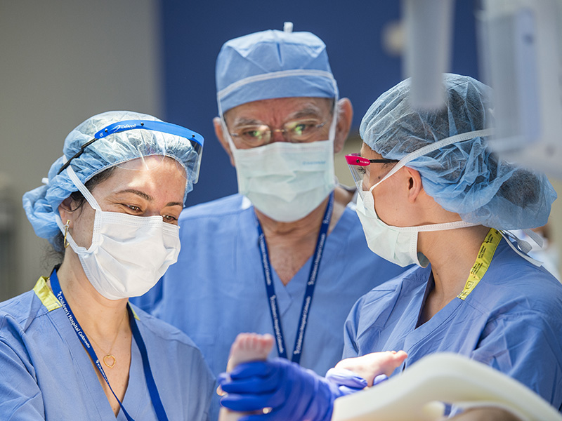 Dr. Alberto Pena, wearing light blue scrubs, a hair covering and white mask stands at an operating table with two women in matching light blue scrubs holding a baby's feet with blue latex gloves.