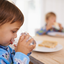 A young child with short brown hair is drinking a glass of water at the breakfast table as a healthy alternative to sugar sweetened beverages like juice.