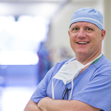 A male doctor wearing blue scrubs has his arms crossed and he's smiling at the camera.