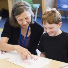 A woman sits with a young boy at a table, examining one of his creative drawings.