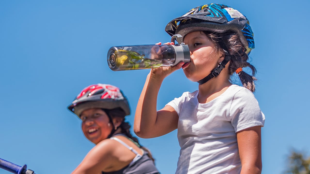A girl wearing a bike helmet drinks water from a water bottle.