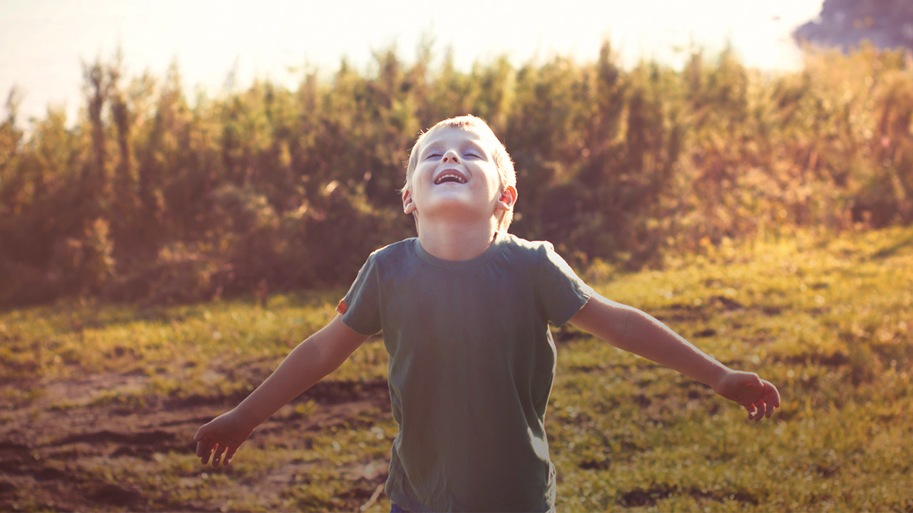 A school-age boy wearing a short sleeve gray shirt stands in a field with tall grasses behind him while the sun shines down. He is looking up smiling with his eyes closed and his arms out to the side.