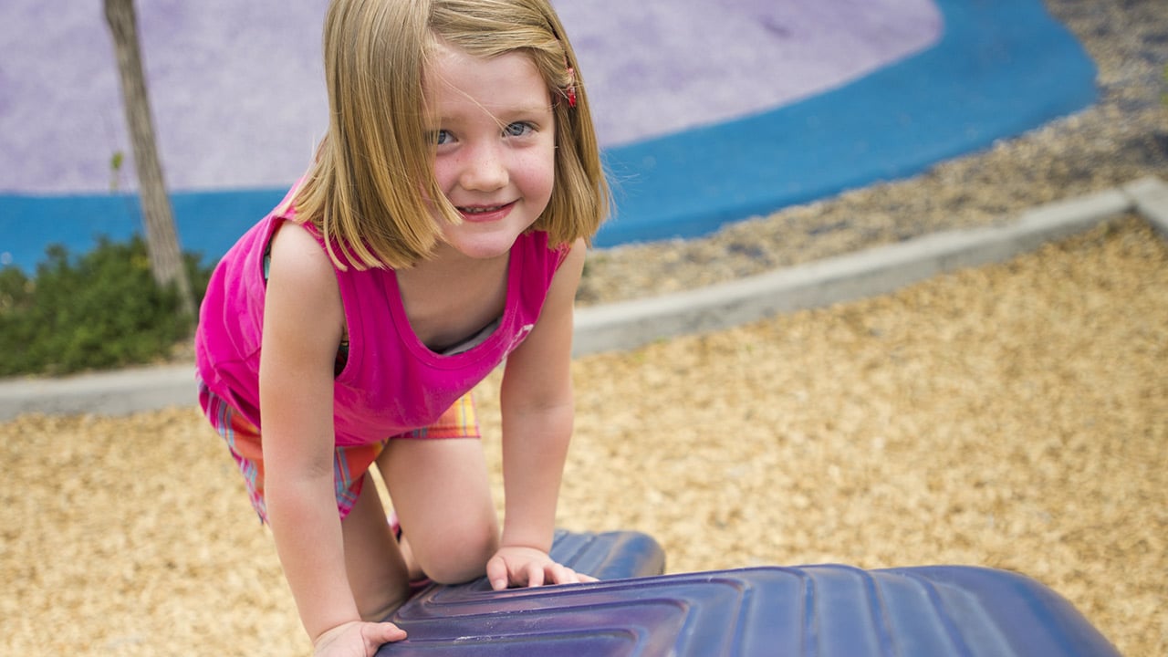 A girl wearing a pink tank top and plaid shorts kneels on a blue wobbly balance beam at the park playground that has wood chips on the ground.