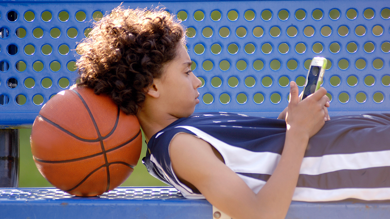 A kid lays on a bench with a basketball while using a cell phone.