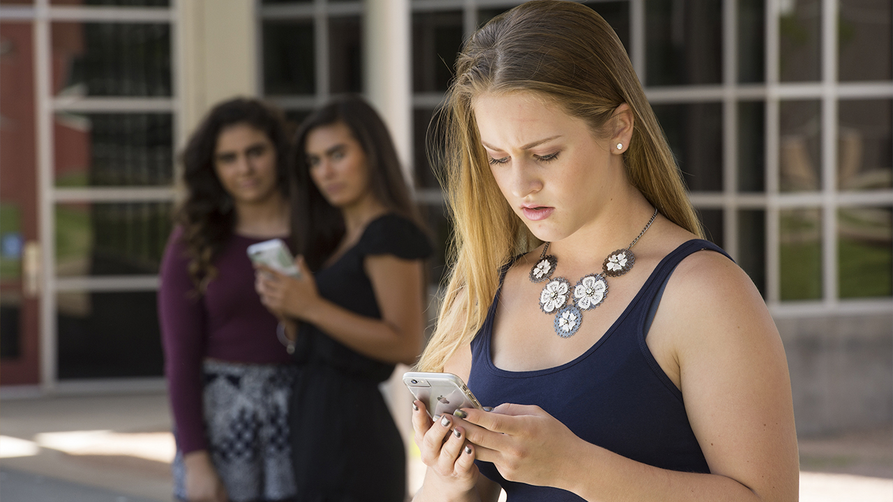 A teenage girl with long hair looks at her phone while two other girls watch from the background.