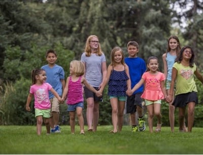 A group of nine kids ranging from toddler to teenager walk in a park holding hands.
