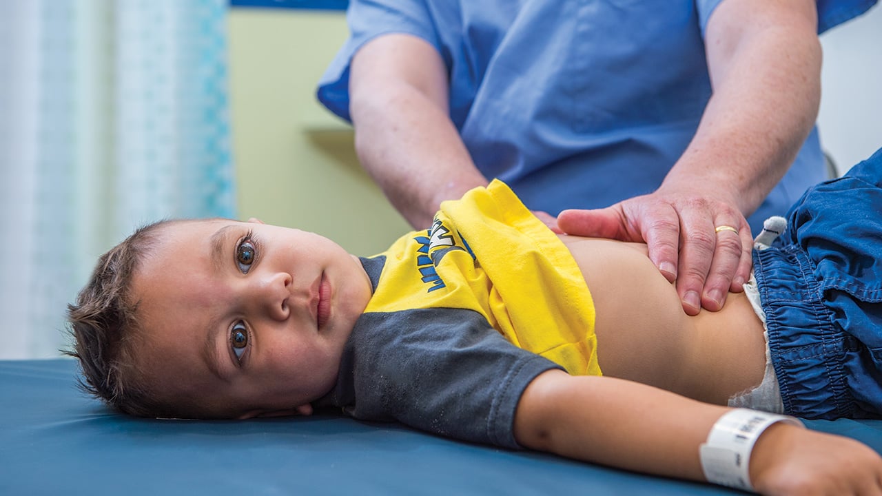 A toddler boy lies on a table while a doctor examines his stomach.