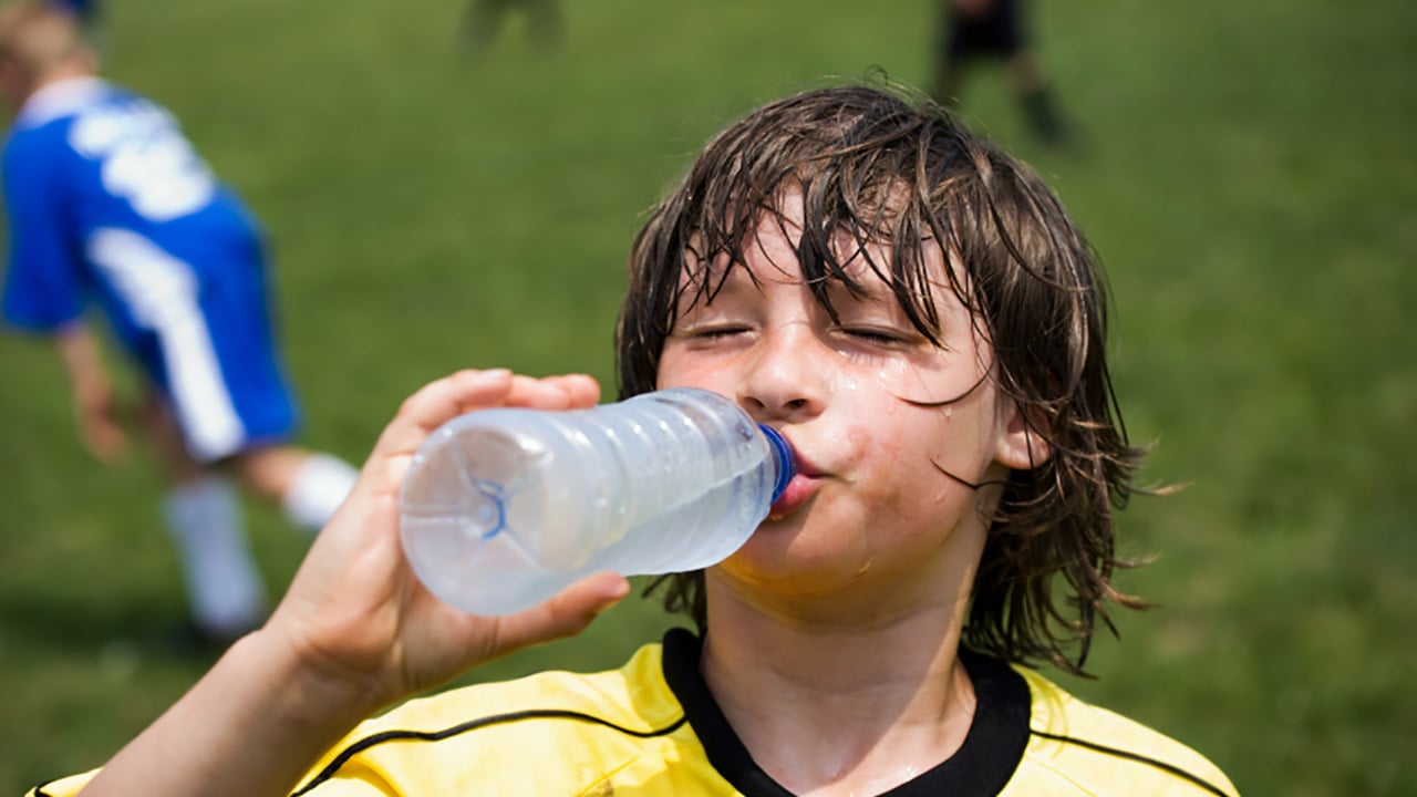 A close-up of a boy with long brown hair and wearing a yellow soccer jersey with black stripe drinking out of a clear water bottle at the soccer field