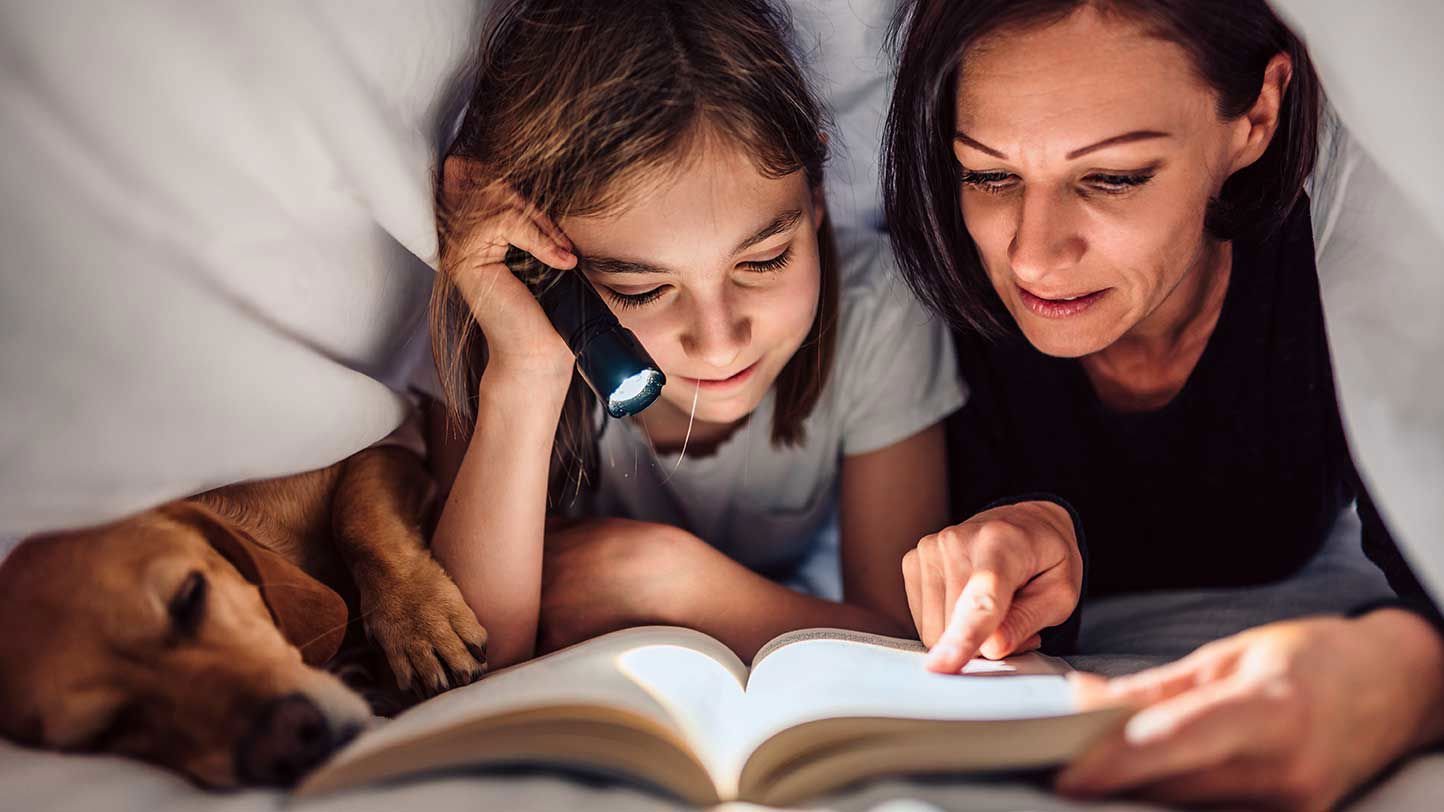 A mom and daughter cuddle with their dog while reading a book under the covers