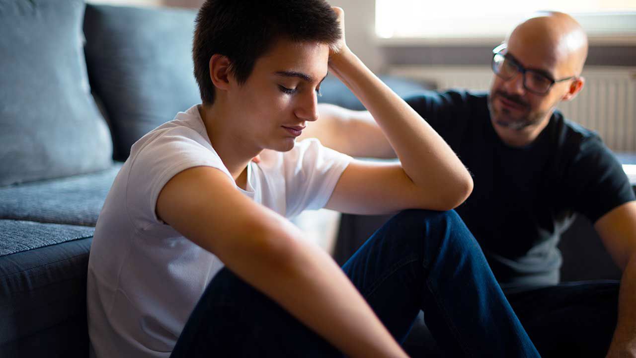 A teen boy and his father sit on the floor in front of a couch. The teen looks upset and his dad is showing support and concern.