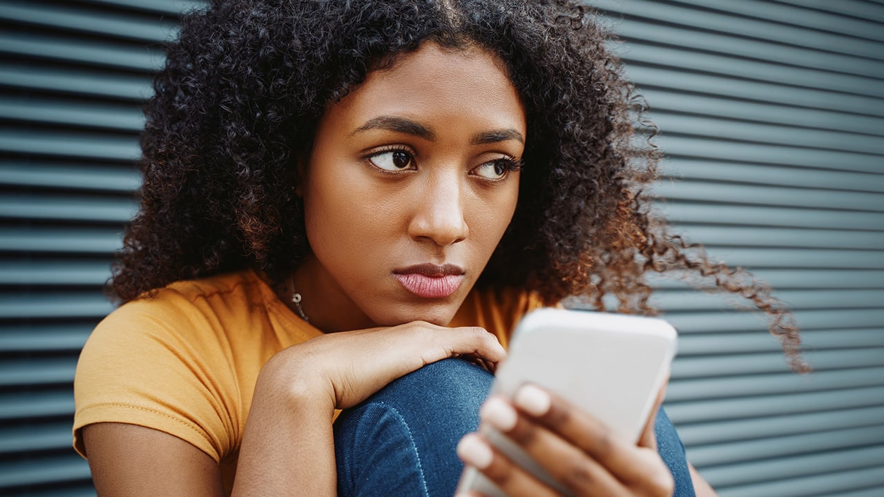 A teenager looks off to the side as she sits with her knees pulled up to her chest and her phone in her hand.