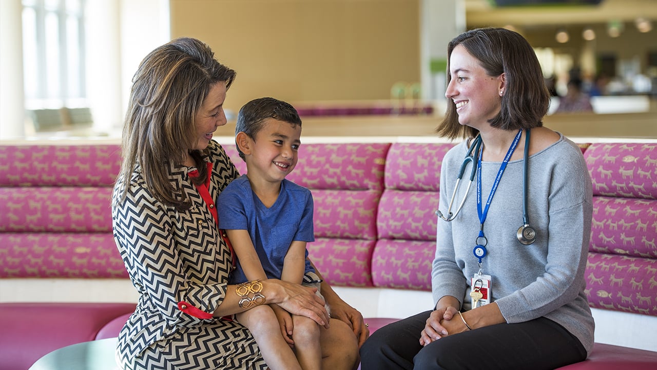 A boy sits on his mom's lap while talking to a doctor in the lobby.