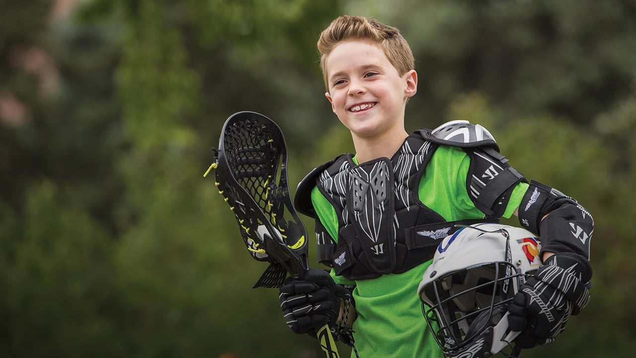 A boy with light brown hair is wearing a neon green athletic shirt with black lacrosse pads over it and black gloves while holding a black lacrosse stick and white lacrosse helmet.