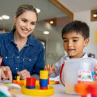 A woman watches as a young child work with bright-colored blocks at a table.