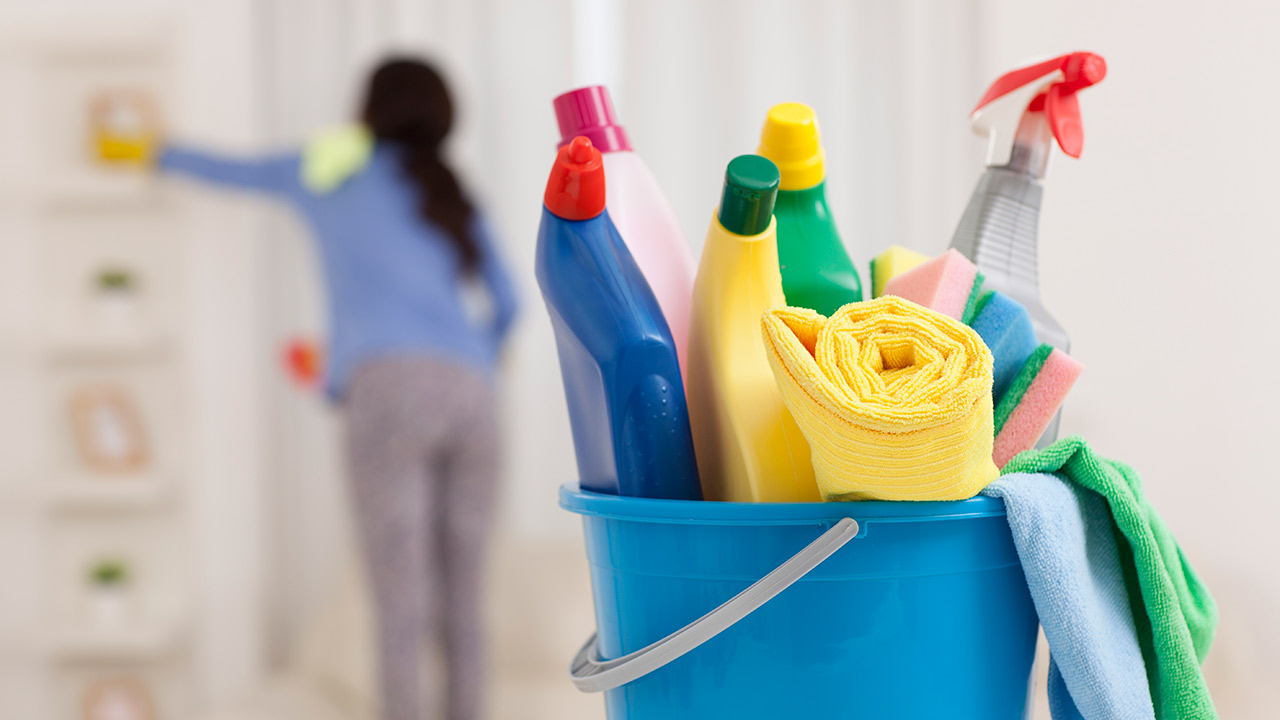 A closeup of a blue bucket holding multiple cleaning bottles and a yellow towel; and out of focus in the background is a woman with long dark hair and wearing a blue shirt while washing a wall.
