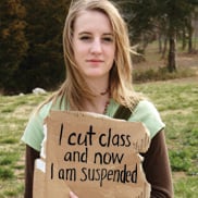 A girl with long hair and wearing a short sleeve light green t-shirt over a long sleeve dark brown t-shirt holds a cardboard sign that says "I cut class and now I am suspended".