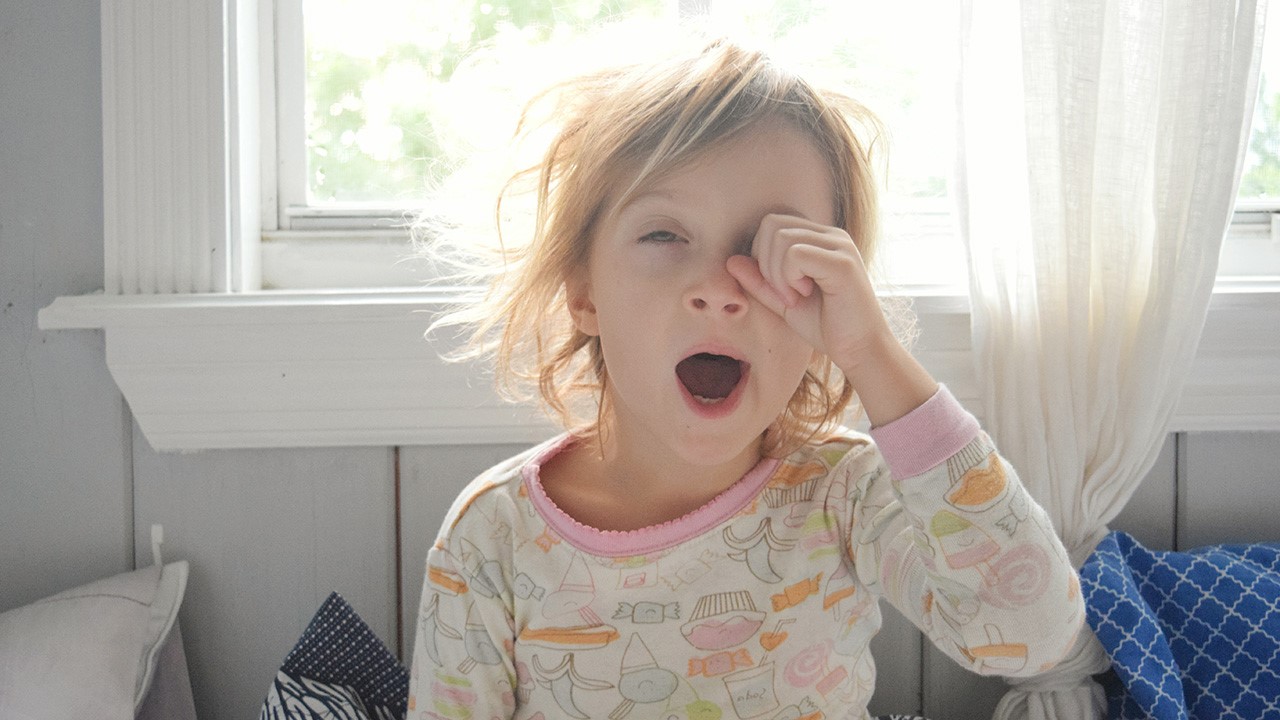 A young girl in pajamas yawning and rubbing her eye.