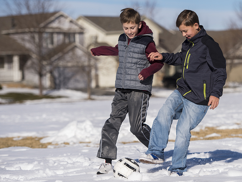 Two boys play soccer in the snow. One boy is wearing a gray vest over a maroon sweatshirt and gray warmup pants. The other boy is wearing a dark gray winter jacket with neon yellow trim and jeans.