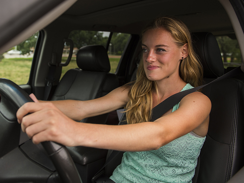 A teenage girl with long blonde hair sits at the wheel of a car.