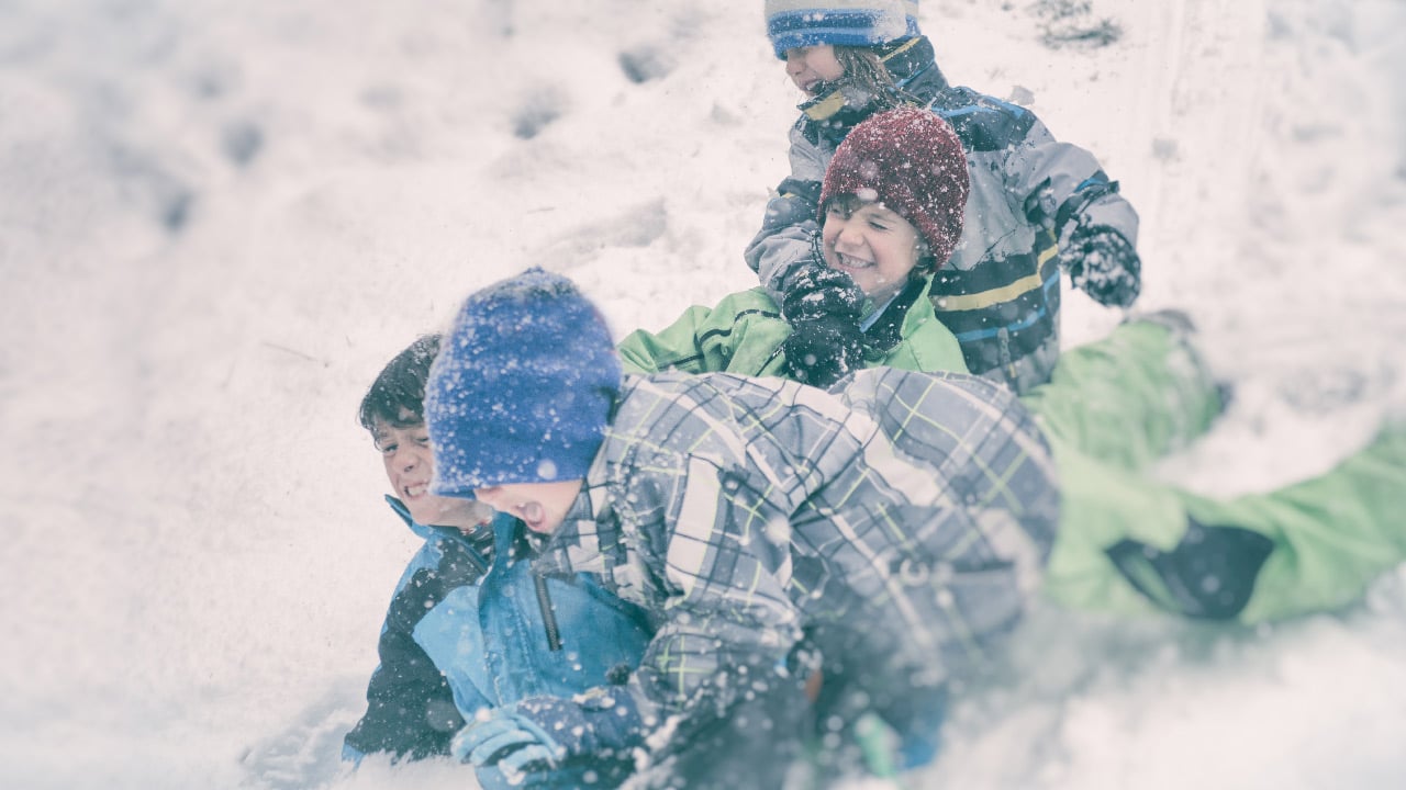 A group of four boys in blue and green snow gear wrestle in the snow.
