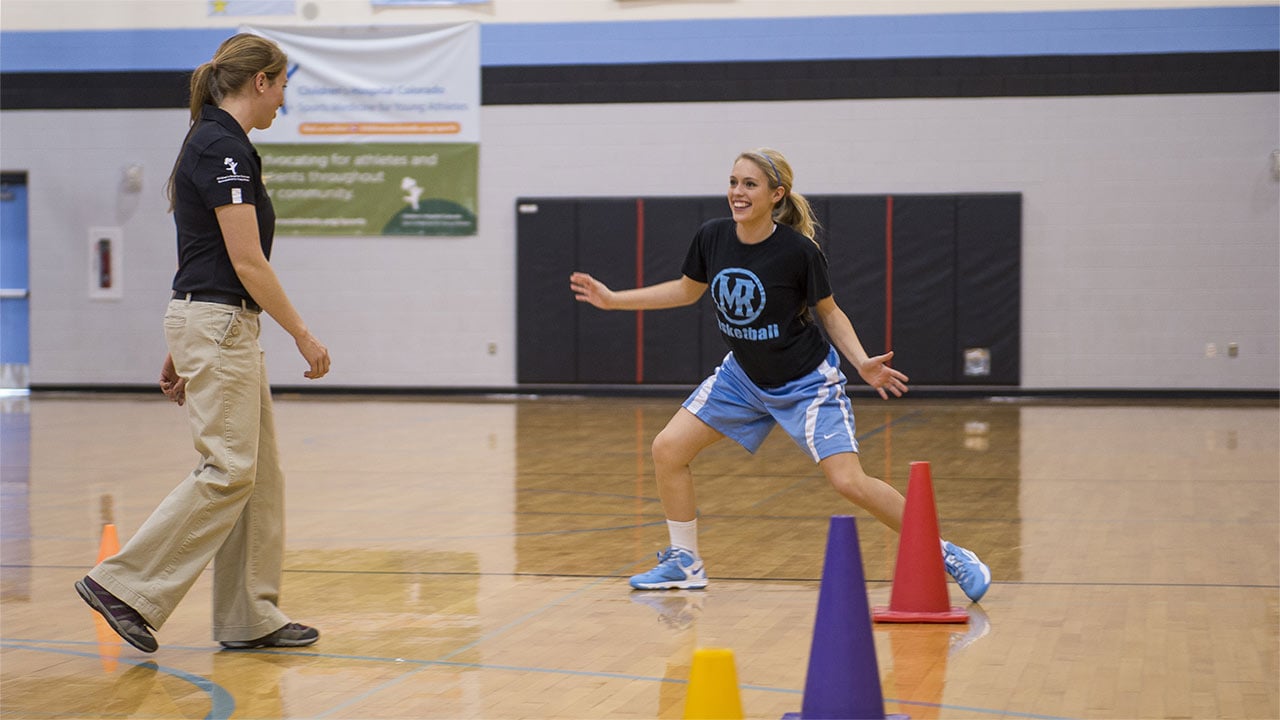 A Sports Medicine Center physical therapist stands in a gym, helping a teenage athlete recover from her sports injury.