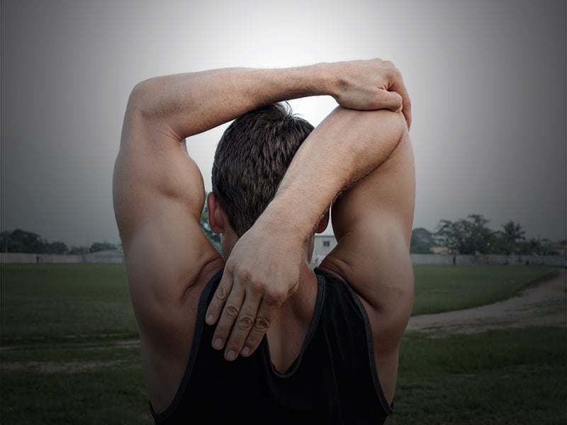 A teenage boy stands in a field doing an overhead tricep stretch.